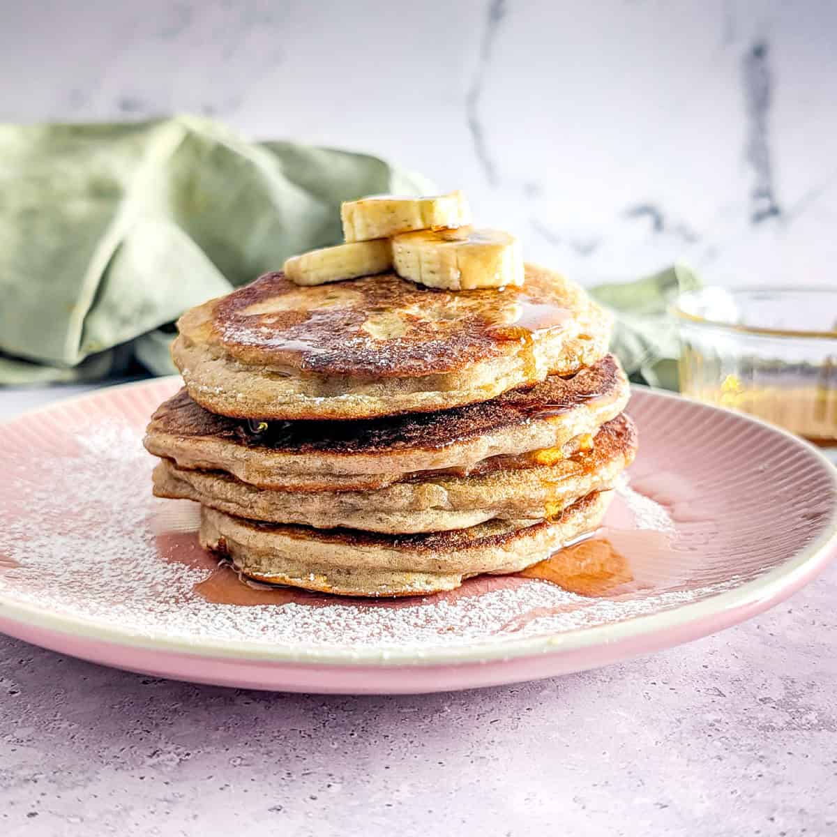 Buckwheat banana pancakes stacked on a pink plate with some maple syrup and sliced bananas.