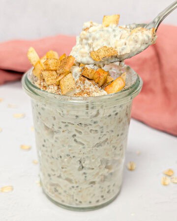Apple cinnamon overnight oats in a glass jar topped with diced apples and cinnamon, with a spoonful lifted.