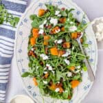 Overhead view of roasted pumpkin chickpea salad with arugula, crumbled feta and yogurt tahini dressing on a white oval serving platter.