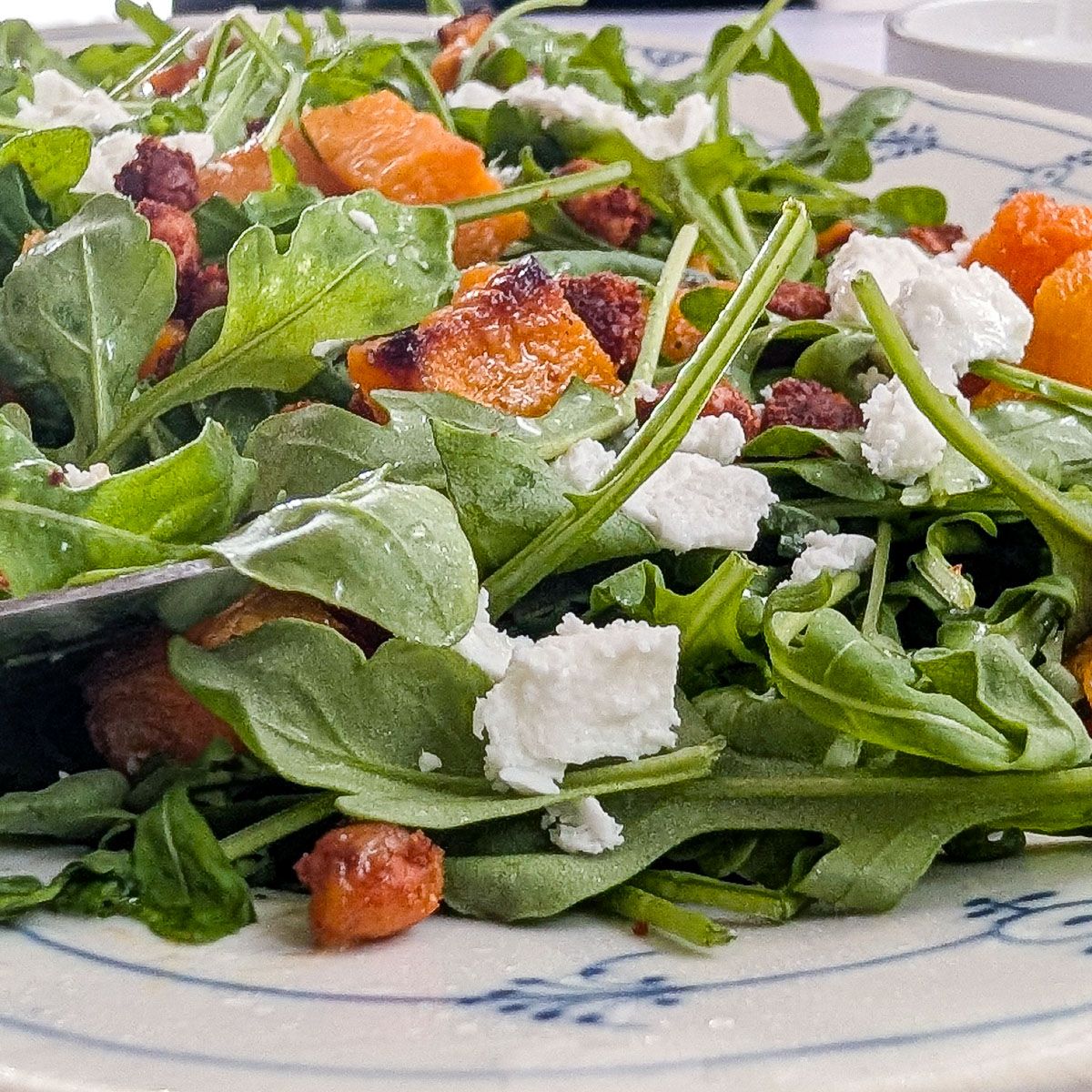 Close-up of roasted pumpkin chickpea salad with arugula, crumbled feta cheese and golden roasted chickpeas on a white plate with blue patterns.