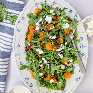 Overhead view of roasted pumpkin chickpea salad with arugula, crumbled feta and yogurt tahini dressing on a white oval serving platter.