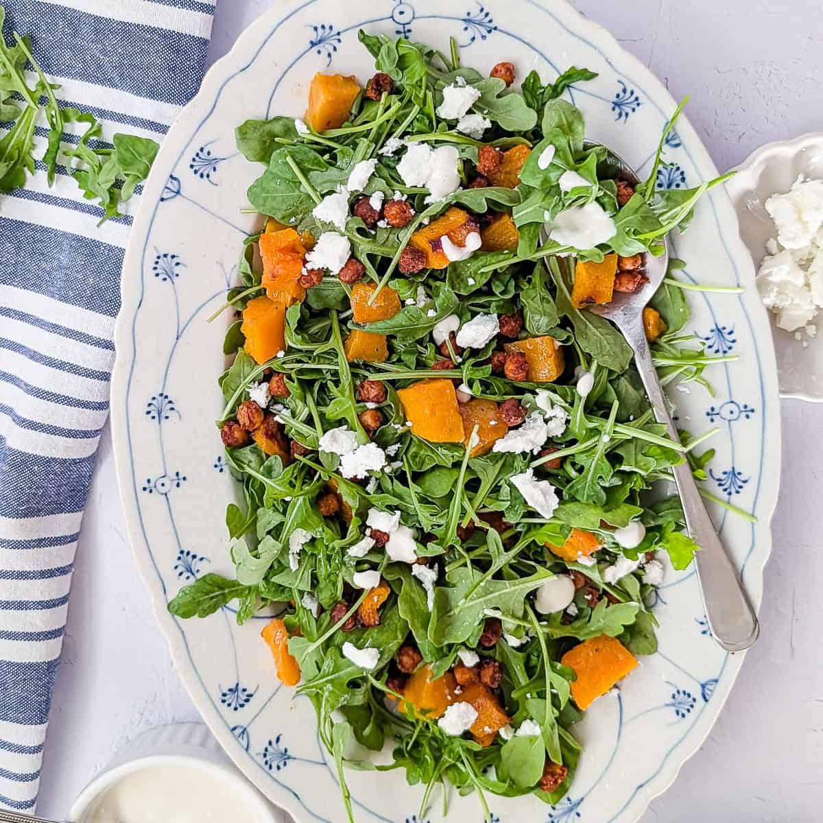 Overhead view of roasted pumpkin chickpea salad with arugula, crumbled feta and yogurt tahini dressing on a white oval serving platter.